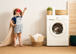kid dressed up in cape and mask next to washing machine basket towels