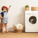 kid dressed up in cape and mask next to washing machine basket towels