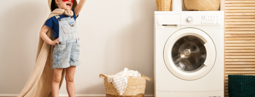 kid dressed up in cape and mask next to washing machine basket towels