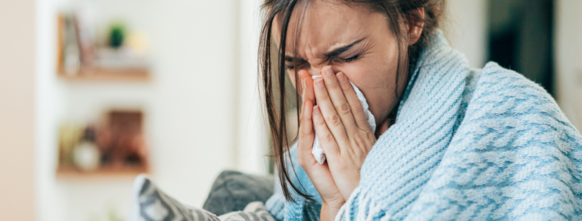 sick woman blowing nose while bundled under blanket.