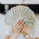 a financially confidence woman holding fanned dollar bills in hands covering face