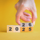 26 ways to save $5 a day. male hand flipping over wooden blocks with year from 2025 to 2026