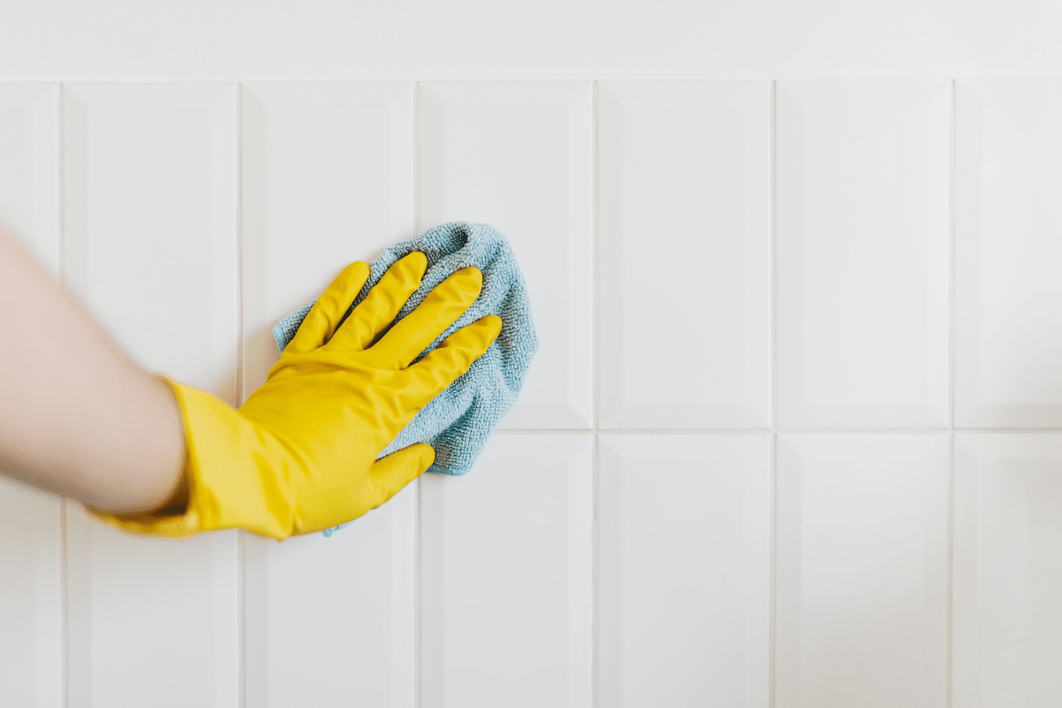 woman's hand in yellow reusable gloves scrubbing white bathroom tile with blue microfiber cloth