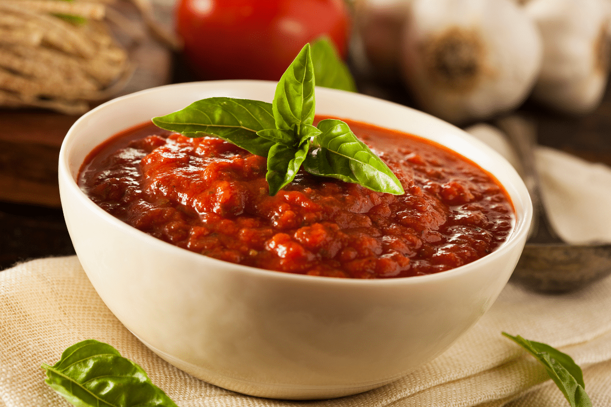 classic homemade marinara sauce recipe in white bowl with fresh basil leaves garlic tomato in background