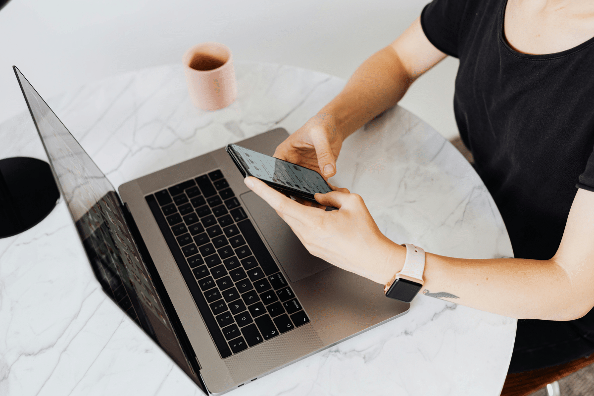 woman sending a text message on phone while sitting behind laptop in kitchen