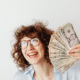 woman holding a fan of money dollar bills smiling moneymaxxing