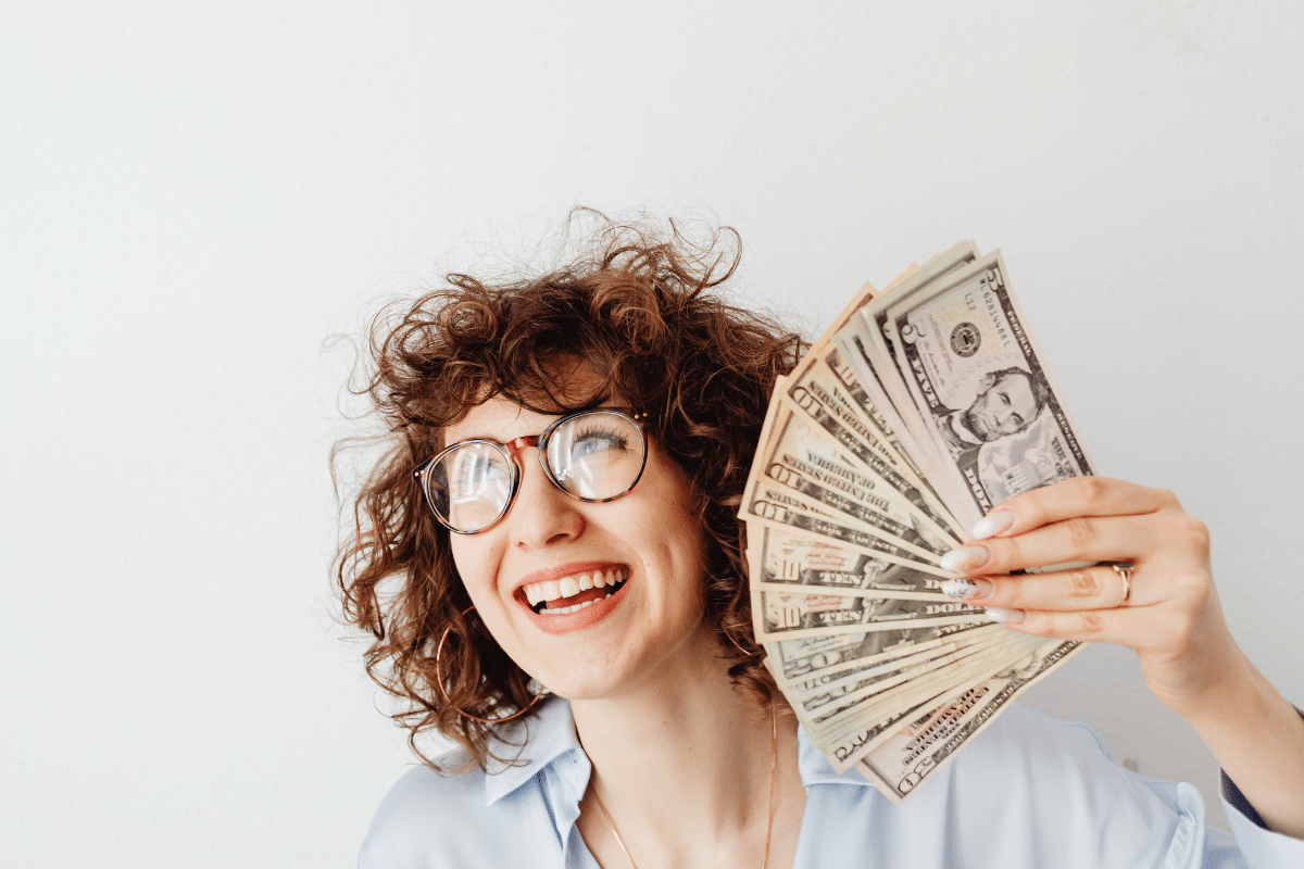 woman holding a fan of money dollar bills smiling moneymaxxing