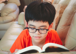 Young asian boy wear glasses reading book and smiling while lying for leisure on sofa in the room with happiness.