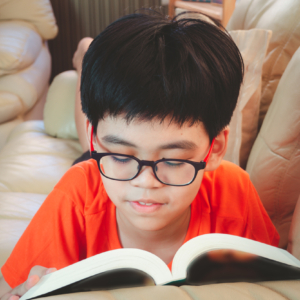 Young asian boy wear glasses reading book and smiling while lying for leisure on sofa in the room with happiness.