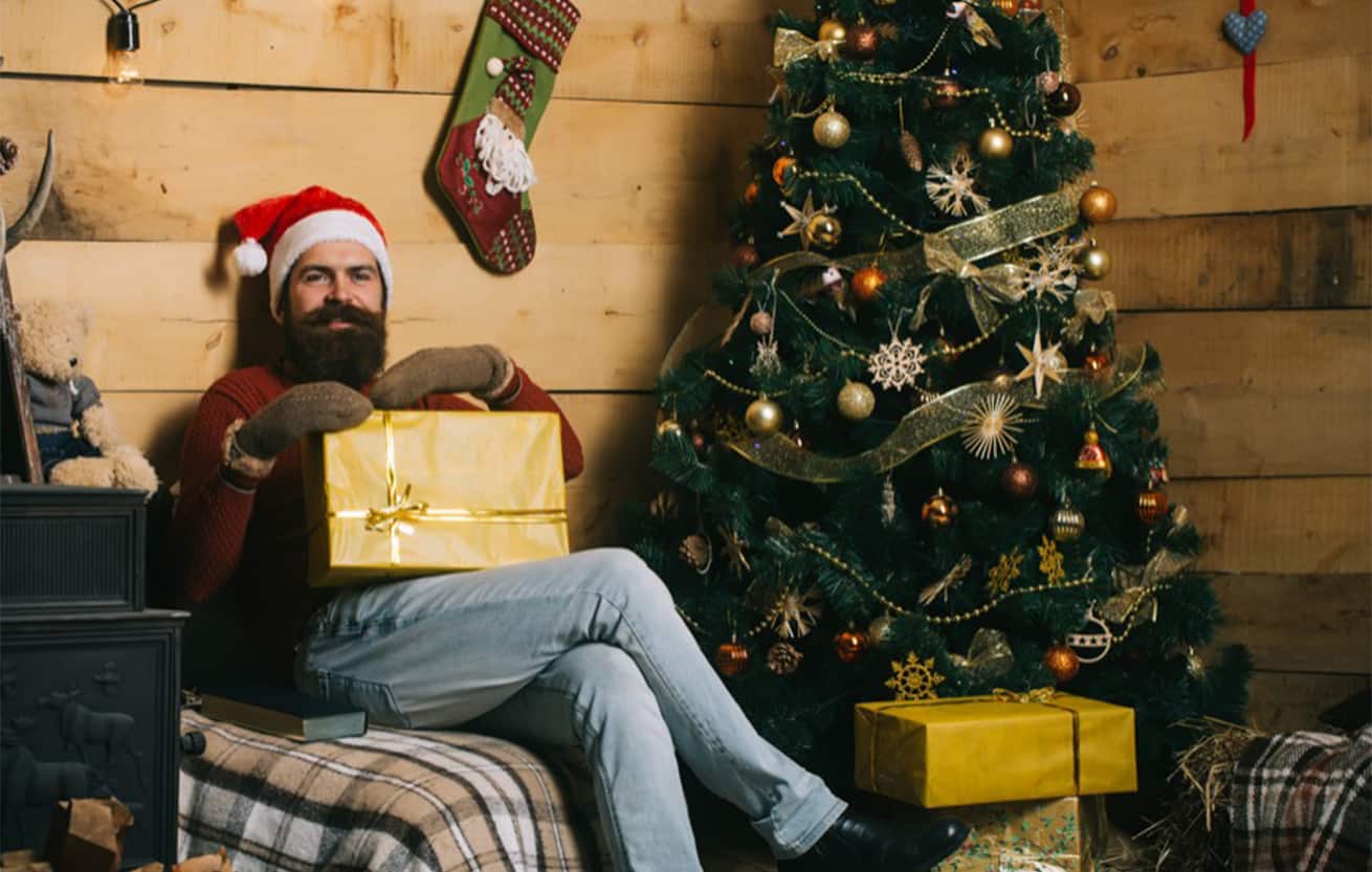 Happy bearded man sitting by Christmas tree with gift in hand
