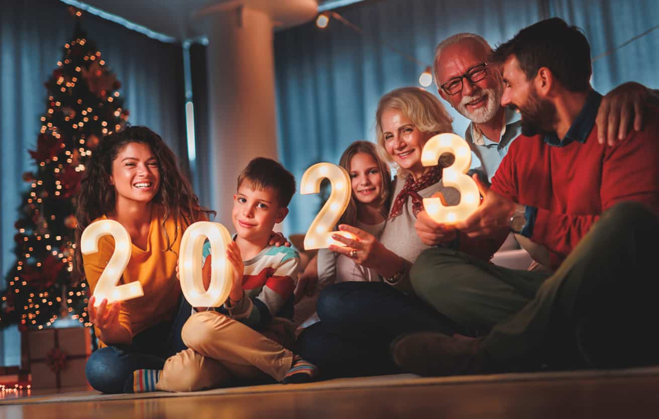 Happy family celebrating New Years Eve at home with kids, sitting by the Christmas tree, holding sparklers and illuminative numbers 2023 representing the upcoming New Year