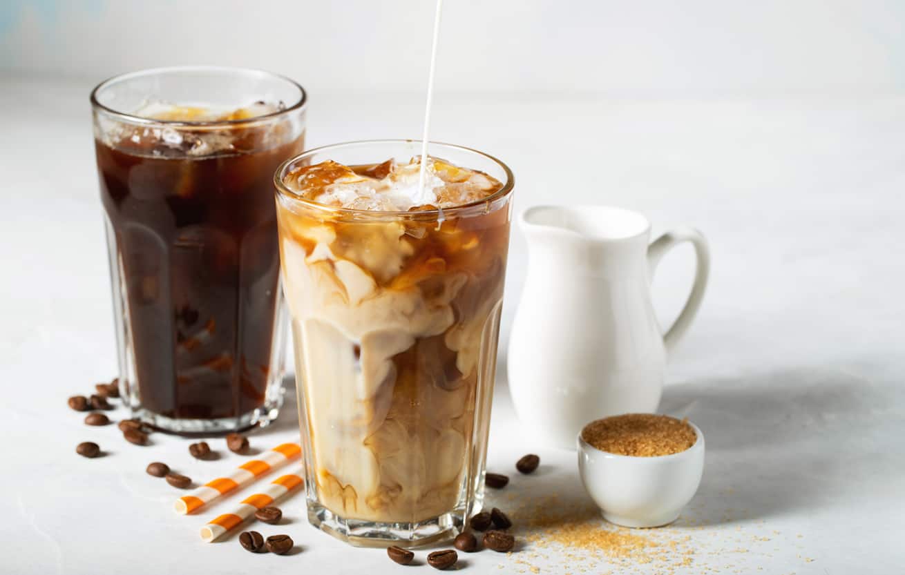 Ice coffee in a tall glass with cream poured over and coffee beans. Cold summer drink on a light blue background.