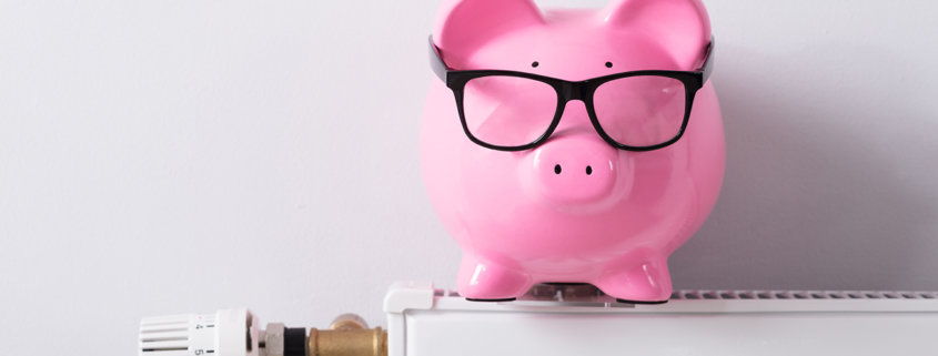 Close-up Of Thermostat And Piggy Bank With Eyeglasses On Radiator Against White Wall