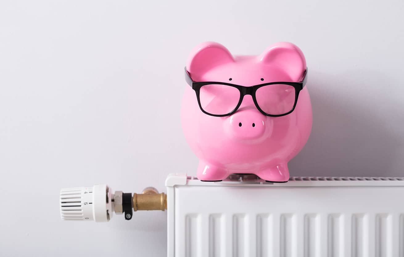 Close-up Of Thermostat And Piggy Bank With Eyeglasses On Radiator Against White Wall