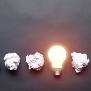 Lightbulb and crumpled white paper balls on black background. Successful solution of problem. Think outside the box. Business motivation with copy space. Genius idea among failing ideas metaphor.