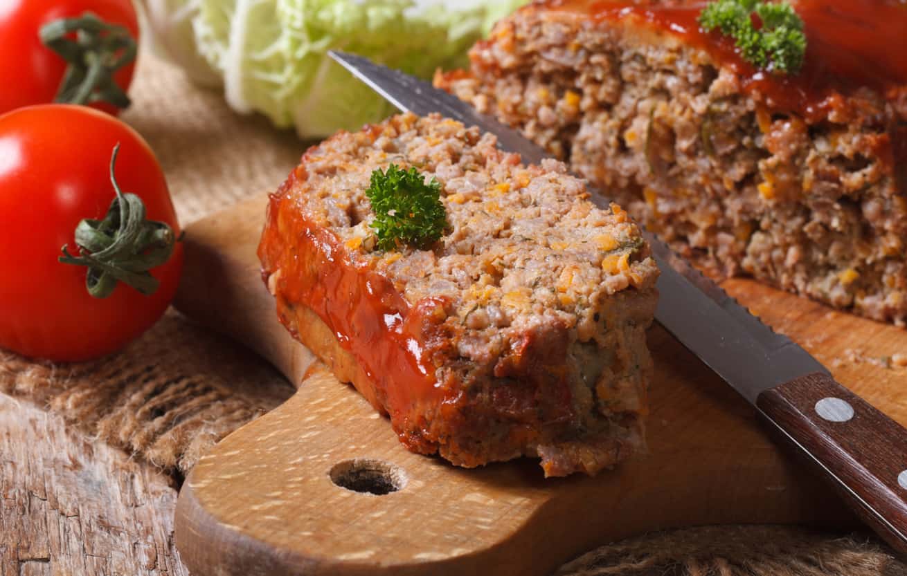 Slice of meatloaf on a cutting board surrounded by vegetables.