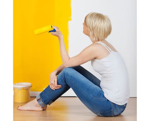 young casual woman with paint roller sitting on floor and looking at half yellow painted wall