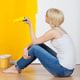 young casual woman with paint roller sitting on floor and looking at half yellow painted wall