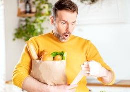 Surprised man looking at store receipt after shopping, holding a paper bag with healthy food. Guy in the kitchen. Real people expression. Inflation concept.