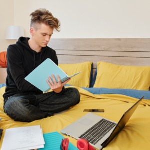 Software engineering student sitting on bed with books, laptop and textbooks around and writing in notebook