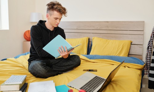 Software engineering student sitting on bed with books, laptop and textbooks around and writing in notebook