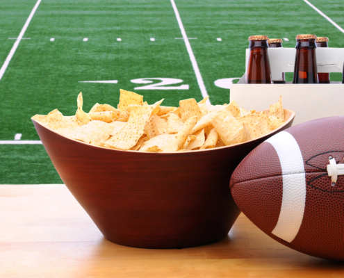 Chips, football and Six Pack of Beer on a table in front of a big screen TV with a Football field.