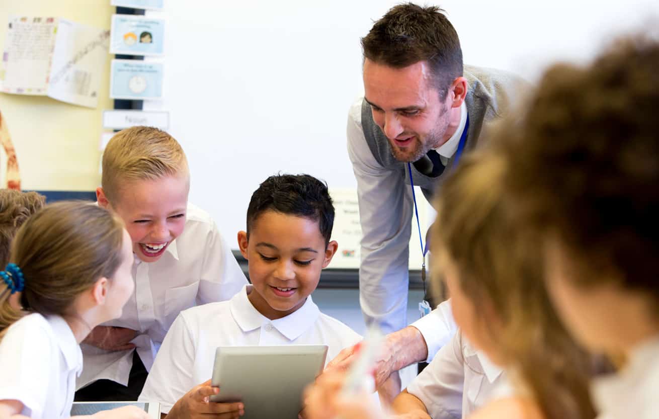 A male teacher sits supervising a group of children who are working on whiteboards and digital tablets