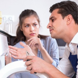 Worried young housewife and repairman near broken washing machine at home kitchen