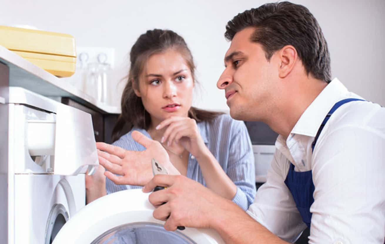 Worried young housewife and repairman near broken washing machine at home kitchen