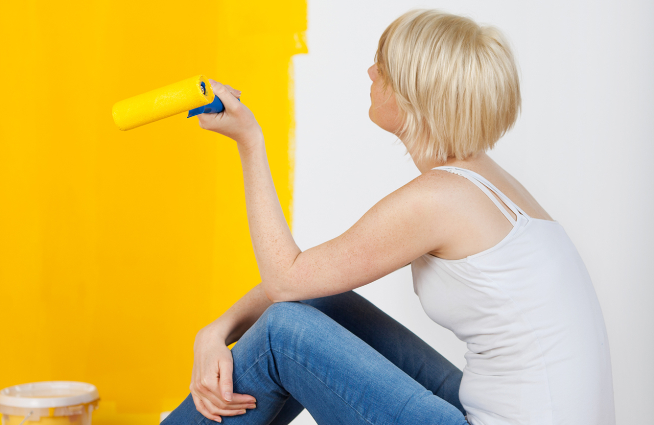 Young casual woman with paint roller sitting on floor and looking at half yellow painted wall