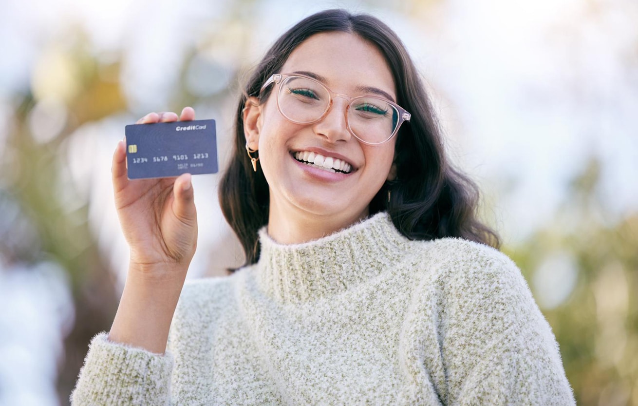 a happy young woman holding up a credit card while standing outside.